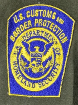 Gregory Bovino wearing a long, structured green wool trench coat with gold buttons and a ceremonial shoulder patch.