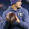 New England Patriots quarterback Drake Maye on the field wearing the slate blue team hoodie and a matching purple-toned beanie with red stars. He is holding a football in both hands, looking toward the side of the frame during a game or practice.