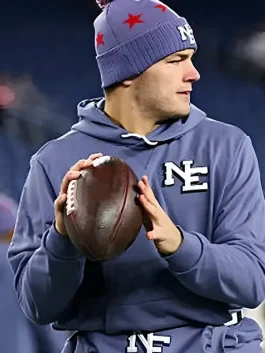 New England Patriots quarterback Drake Maye on the field wearing the slate blue team hoodie and a matching purple-toned beanie with red stars. He is holding a football in both hands, looking toward the side of the frame during a game or practice.