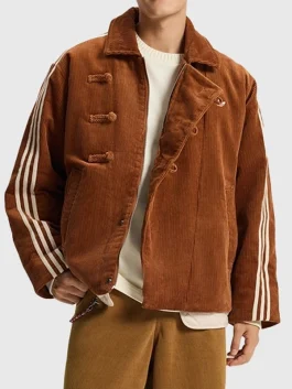 A full-length front view of the unisex Adidas corduroy button jacket worn over a cream sweater and mustard-colored trousers. The design features three prominent corduroy toggle closures on the right side and contrasting white stripes on both sleeves, emphasizing a mix of heritage craftsmanship and modern streetwear.