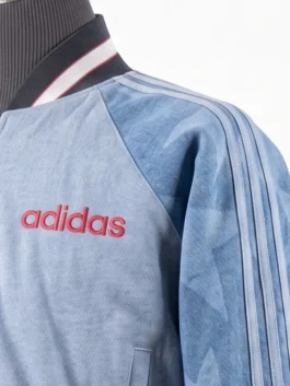 Close-up of the red embroidered Adidas logo on the upper chest of a light blue denim-textured bomber jacket with a striped collar.