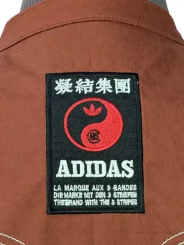 Close-up view of the black rectangular collaboration patch featuring a yin-yang Trefoil logo on the back of the brown Adidas CLOT jacket.
