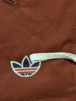 Macro close-up of the tri-color embroidered Adidas Trefoil logo and white side drawcord adjustor on the brown CLOT jacket.
