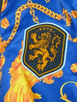 Close-up macro photograph of the KNVB shield emblem on the Netherlands Anthem Jacket, featuring a finely embroidered gold Dutch lion rampant inside a black crest bordered in white.