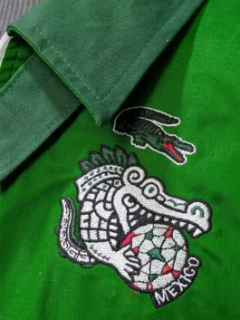 Close-up of embroidered patches on a green jacket featuring a classic dark green crocodile emblem positioned right above a larger Aztec-styled crocodile biting a red, white, and green soccer ball with Mexico text.